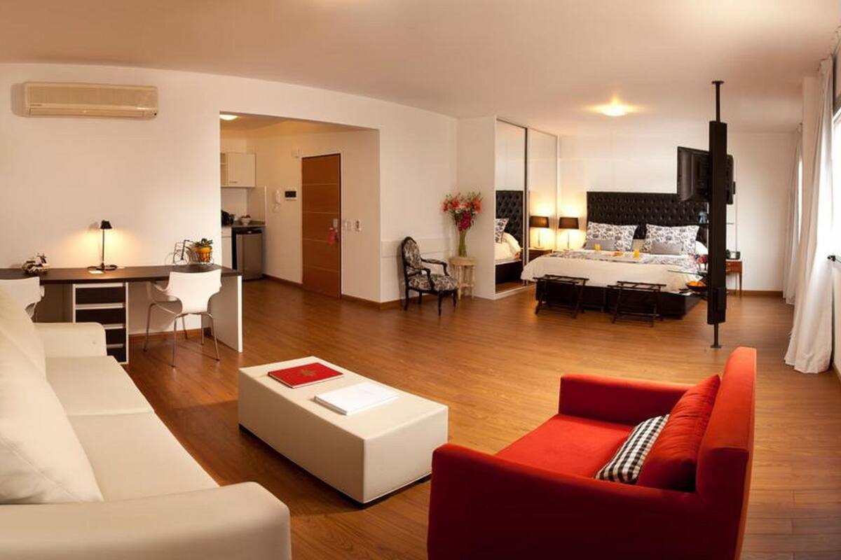 A modern living room with a sofa, a red armchair, a dining table, and a kitchen in the background.