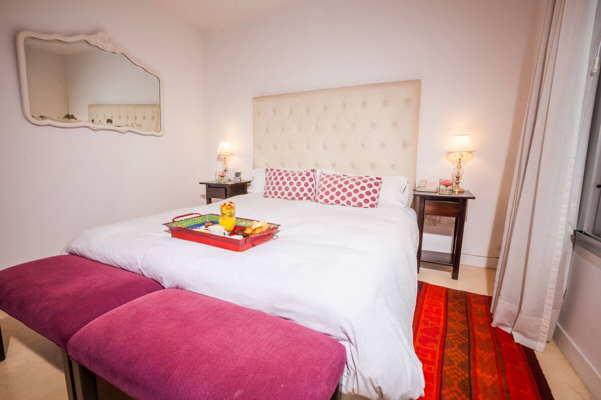 A bedroom with a bed, a bench, two bedside tables with lamps, a mirror, and a window with curtains.