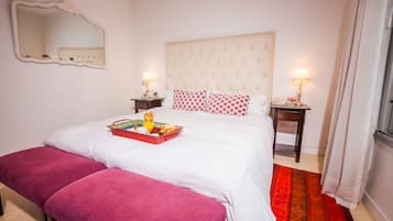 Una camera da letto con un letto, una panca, due comodini con lampade, uno specchio e una finestra con tende.