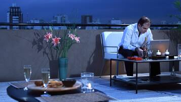 A man seated at a table with a laptop, a candle, and a glass of wine on a balcony overlooking a cityscape.