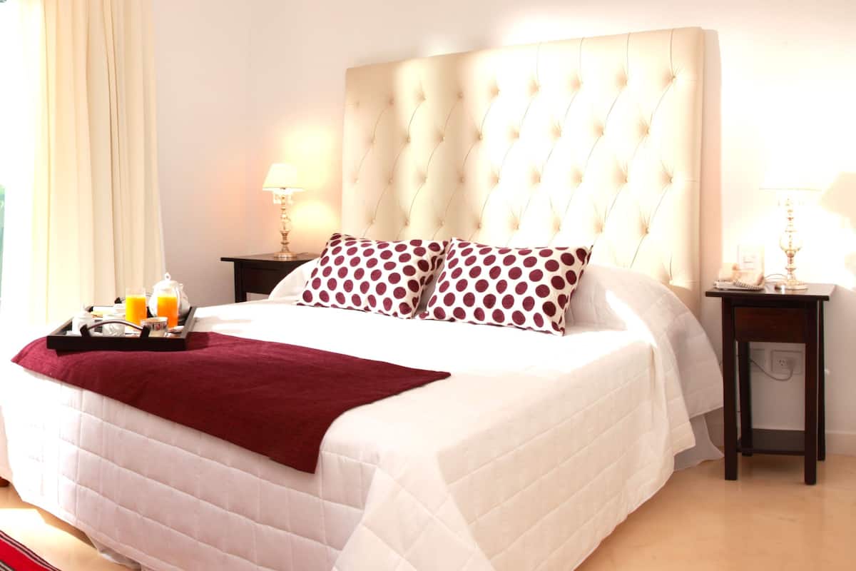 A neatly made bed with a tufted headboard, a red throw blanket, and two patterned pillows. There is a bedside table with a lamp and a tray with a glass of orange juice.