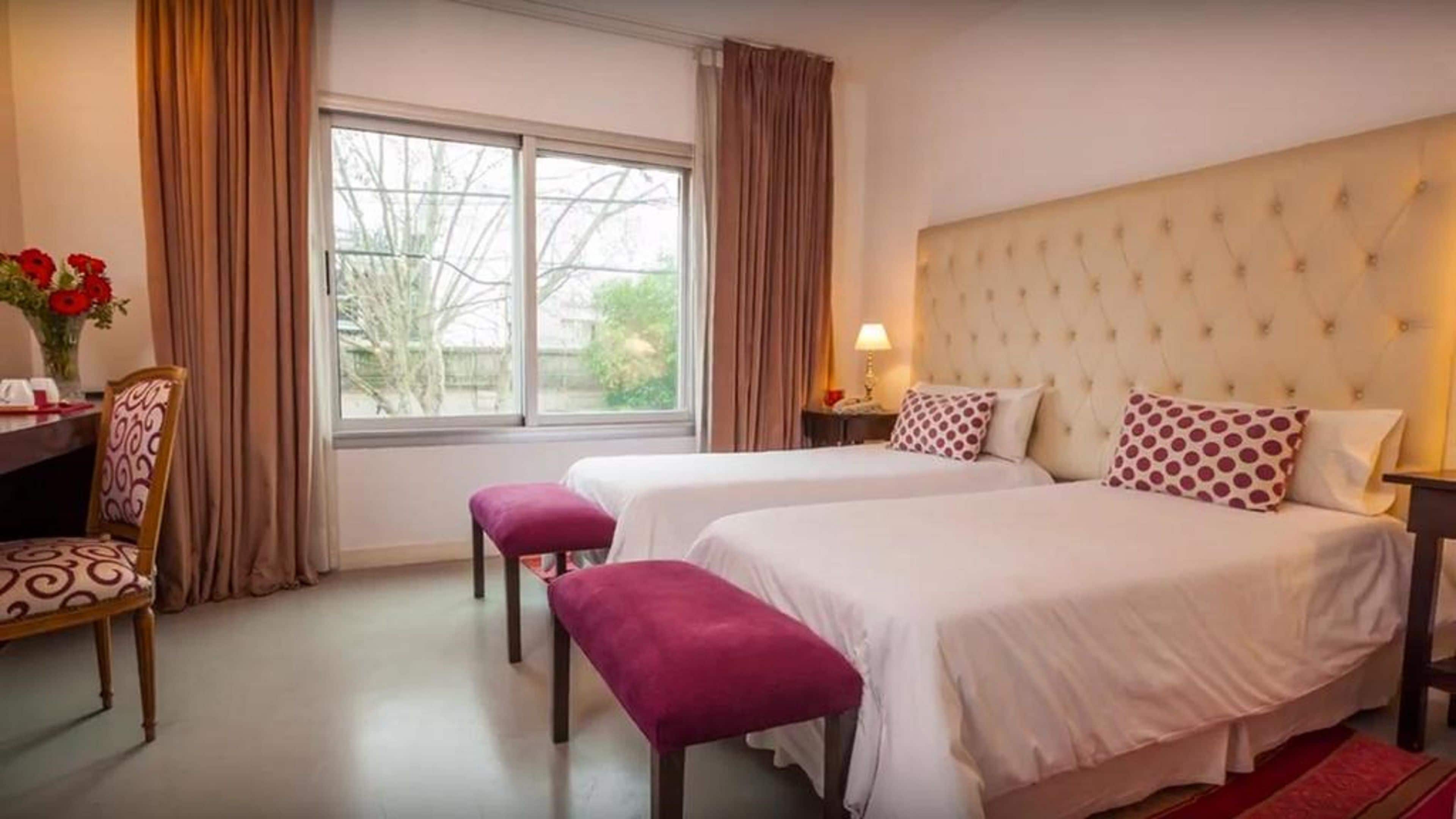 A hotel room with two beds, a desk with a chair, a window with curtains, and a view of trees outside.