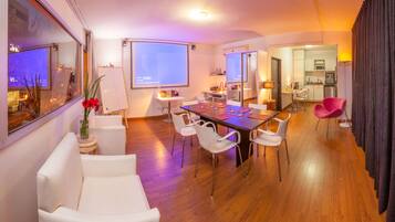 Un salon moderne avec un plancher en bois, des meubles blancs, un écran de projection et un coin repas.