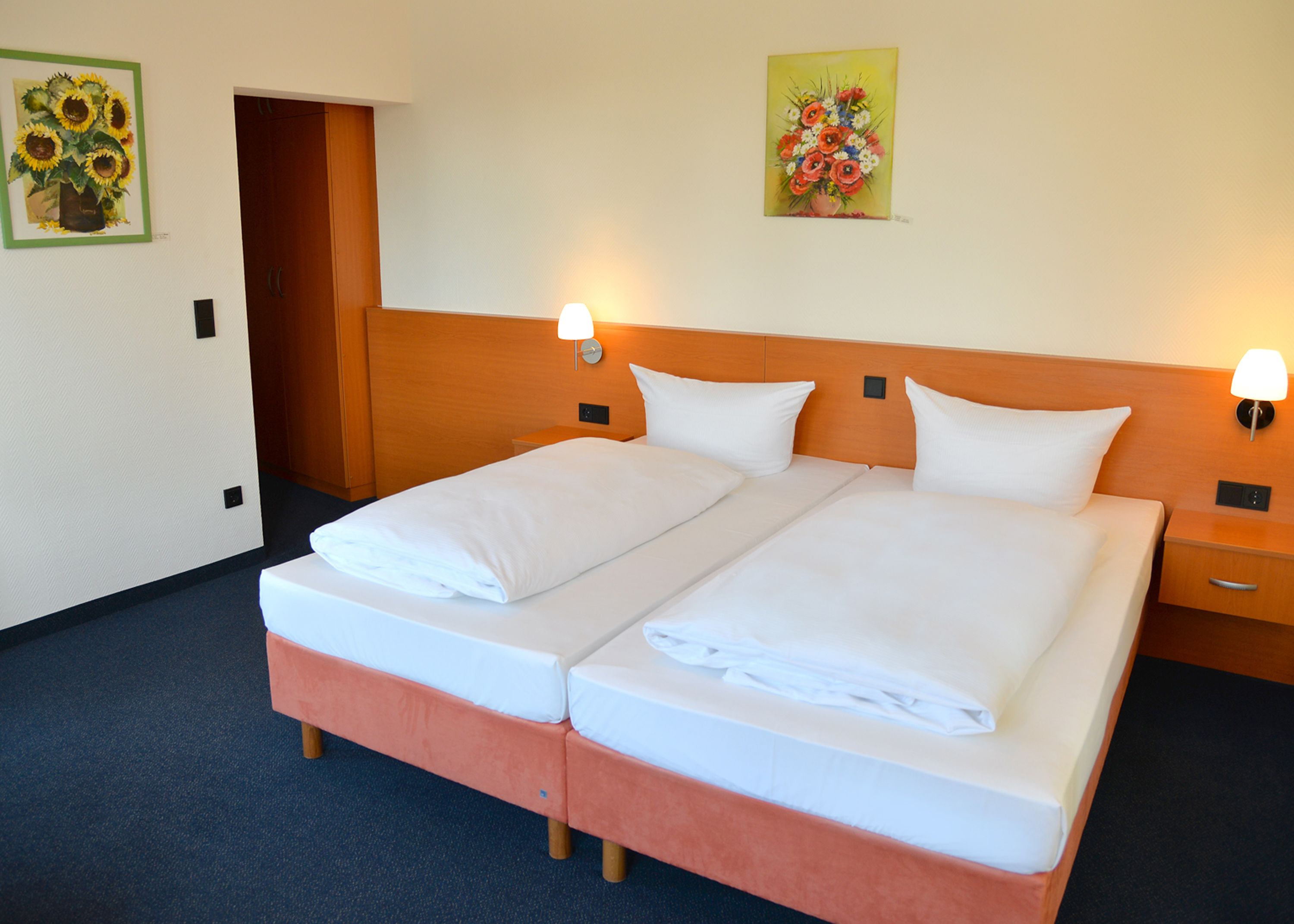 Cama doble con ropa de cama blanca, dos mesitas de noche con lámparas y dos cuadros en las paredes.
