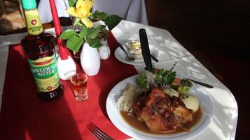 Una mesa de comedor con una botella de Spreewald Bitter, una copa pequeña, una vela roja, un florero blanco con flores amarillas y un plato de comida que incluye carne, patatas y verduras.