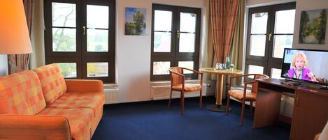 A hotel room with a sofa, a dining table with chairs, a desk with a computer, and large windows with curtains.