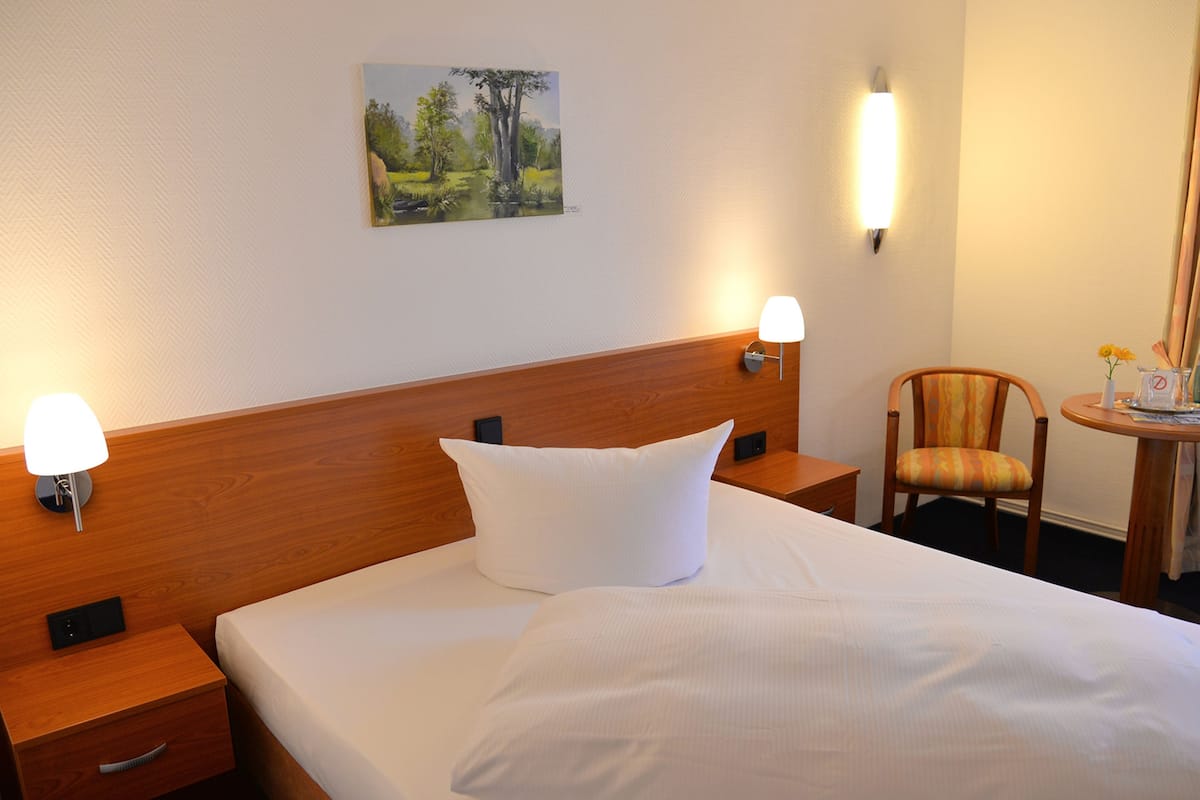 A hotel room with a wooden headboard, a bed with white linens, two bedside lamps, a small table with a chair, and a painting on the wall.