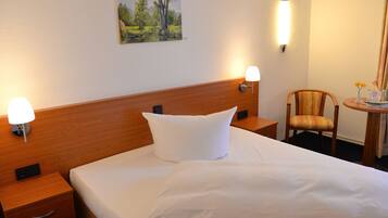 Un cuarto de hotel con cabecera de madera, cama con ropa blanca, dos lámparas de noche, una mesita con una silla y un cuadro en la pared.