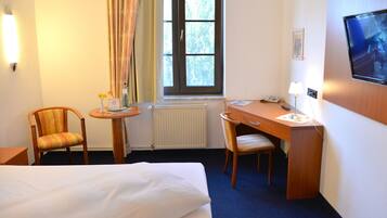 Une chambre d’hôtel avec un lit, un bureau, deux chaises, une petite table sur laquelle est posé un vase de fleurs, une fenêtre avec des rideaux, un radiateur et un téléviseur fixé au mur.