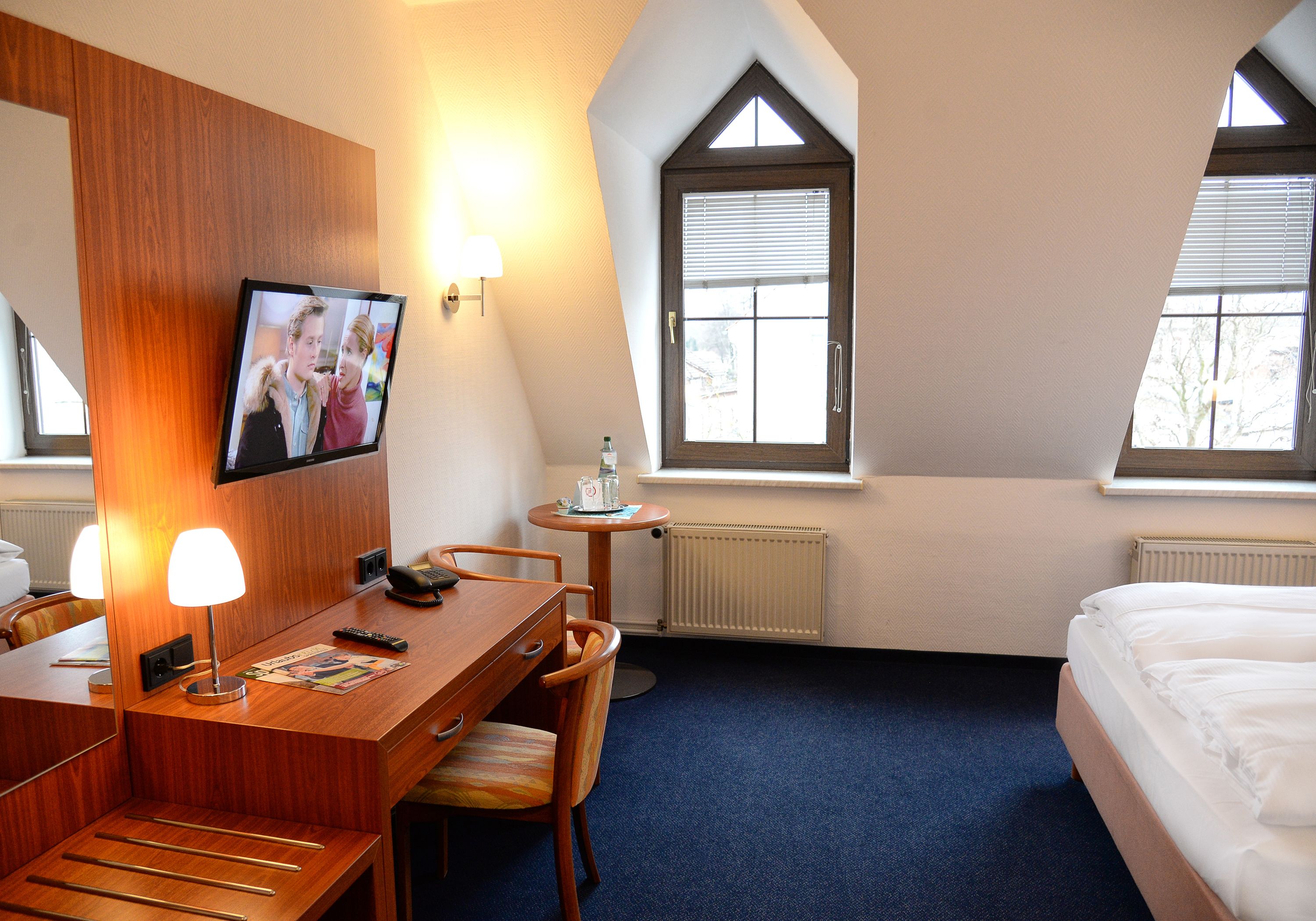 Habitación de hotel con escritorio de madera, cama, televisor montado en la pared y dos ventanas con persianas.