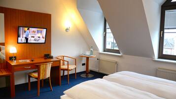 Habitación de hotel con cama, escritorio y silla, televisor montado en la pared, una mesita con otra silla y ventana con persianas.