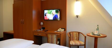 Habitación de hotel con una cama, un escritorio con teléfono, una silla, un televisor montado en la pared y una mesita con una botella.