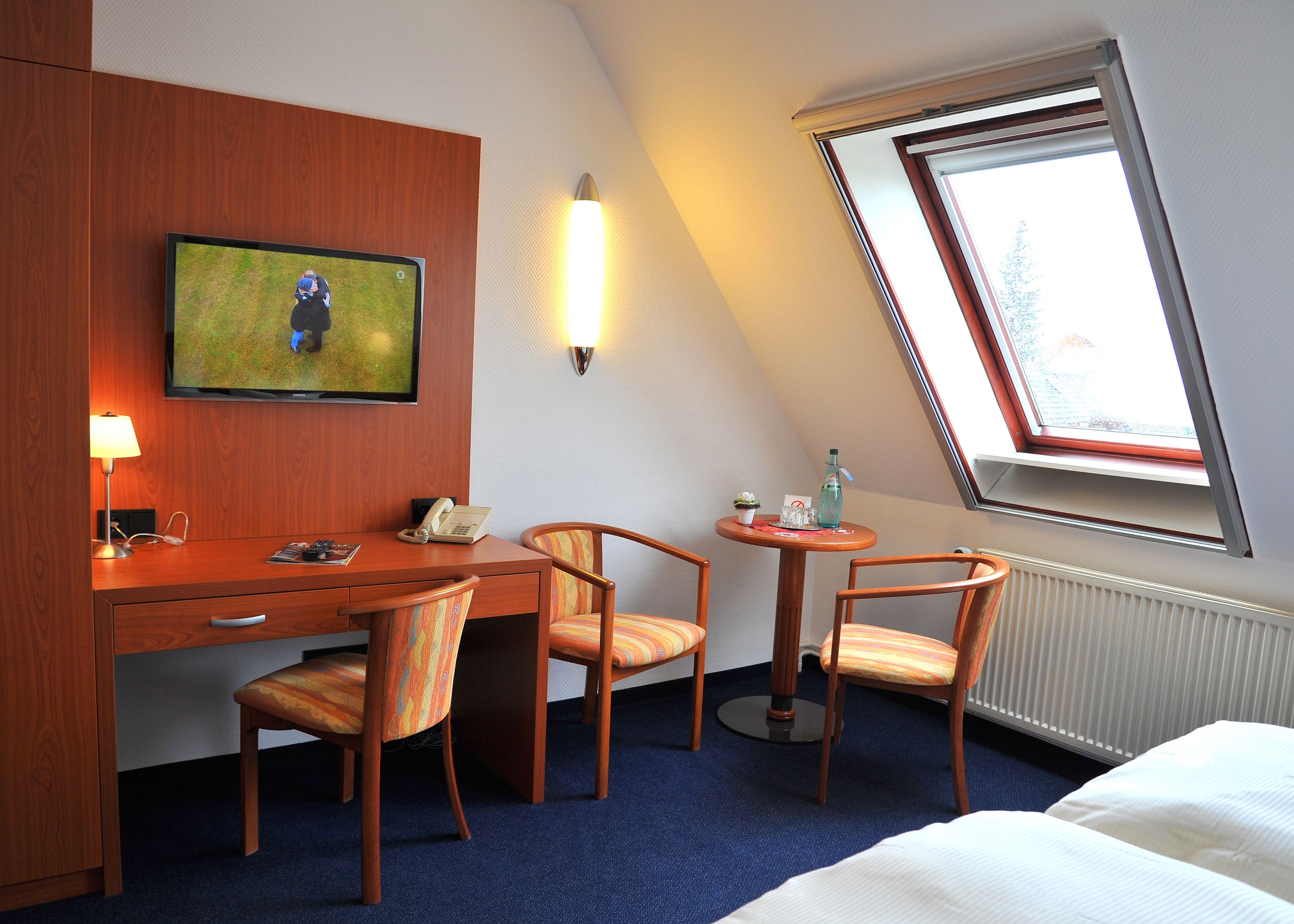 Habitación de hotel con escritorio, dos sillas, televisión, una mesita con una botella y vasos, ventana con vistas, y una cama.