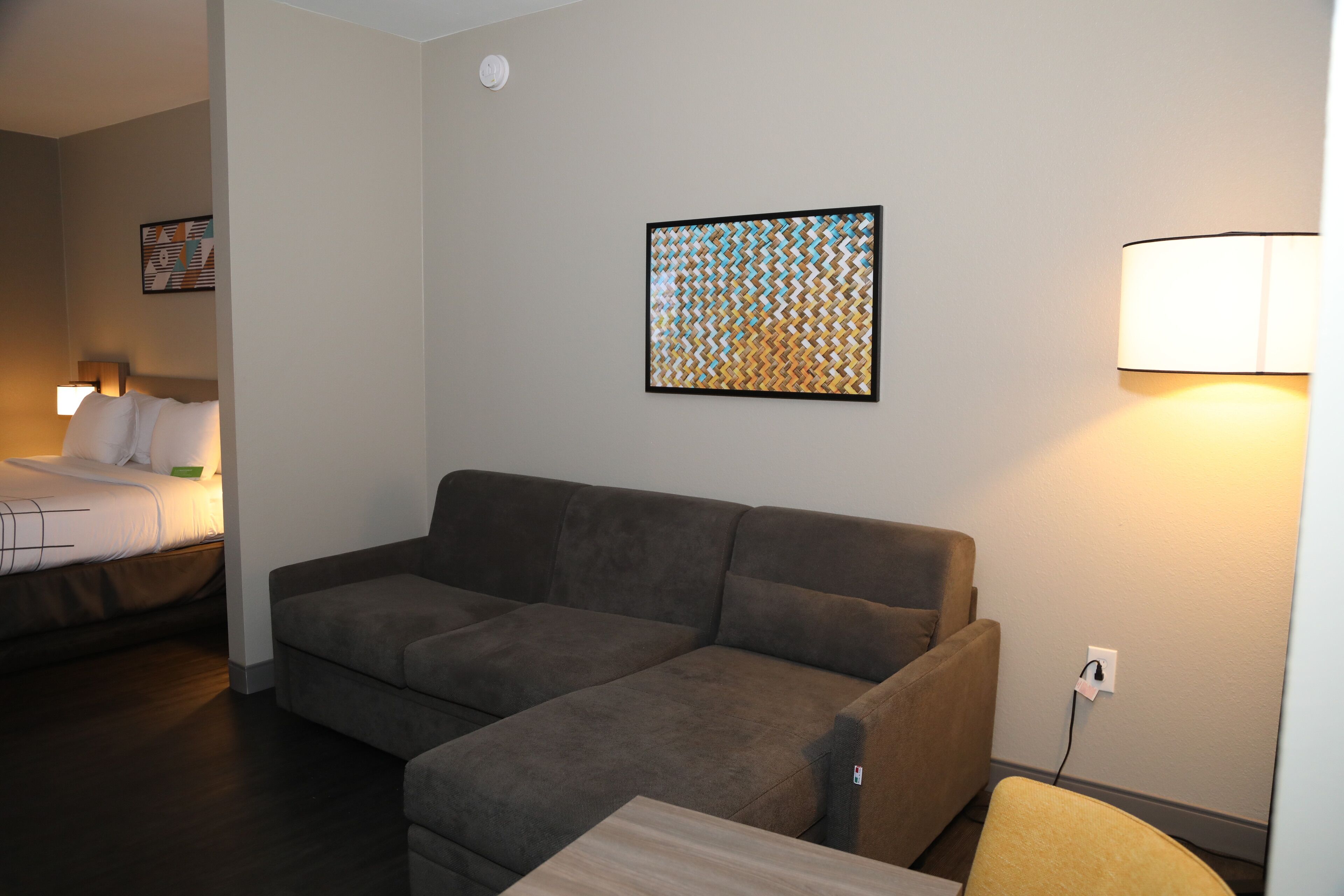 Una habitación de hotel moderna con un sofá marrón, una mesita de madera y una cama con ropa blanca.