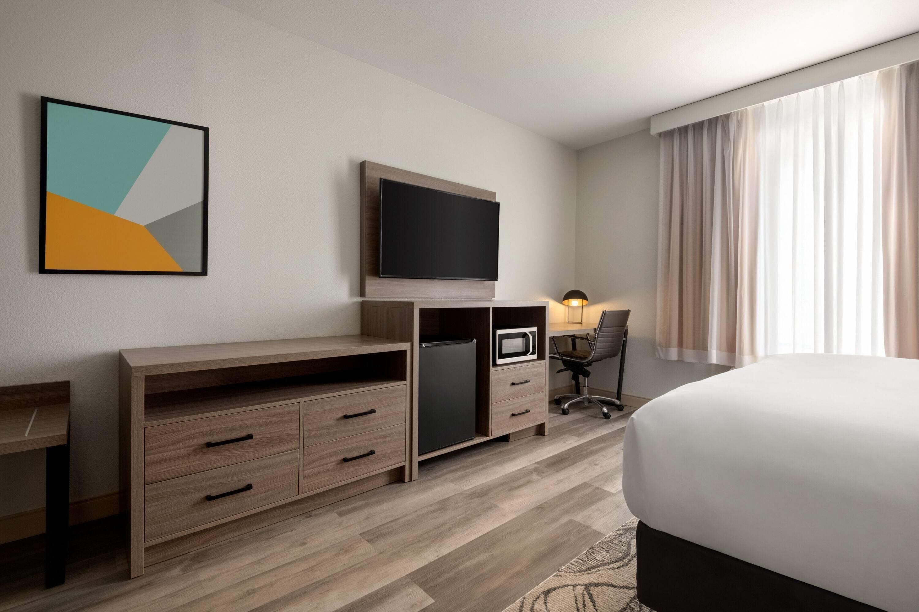 A hotel room with a bed, a desk with drawers, a television, a chair, and a lamp.