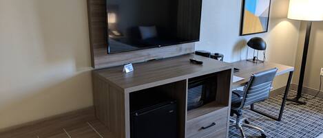 Quarto de hotel com uma escrivaninha de madeira, televisão, luminária e uma cadeira.