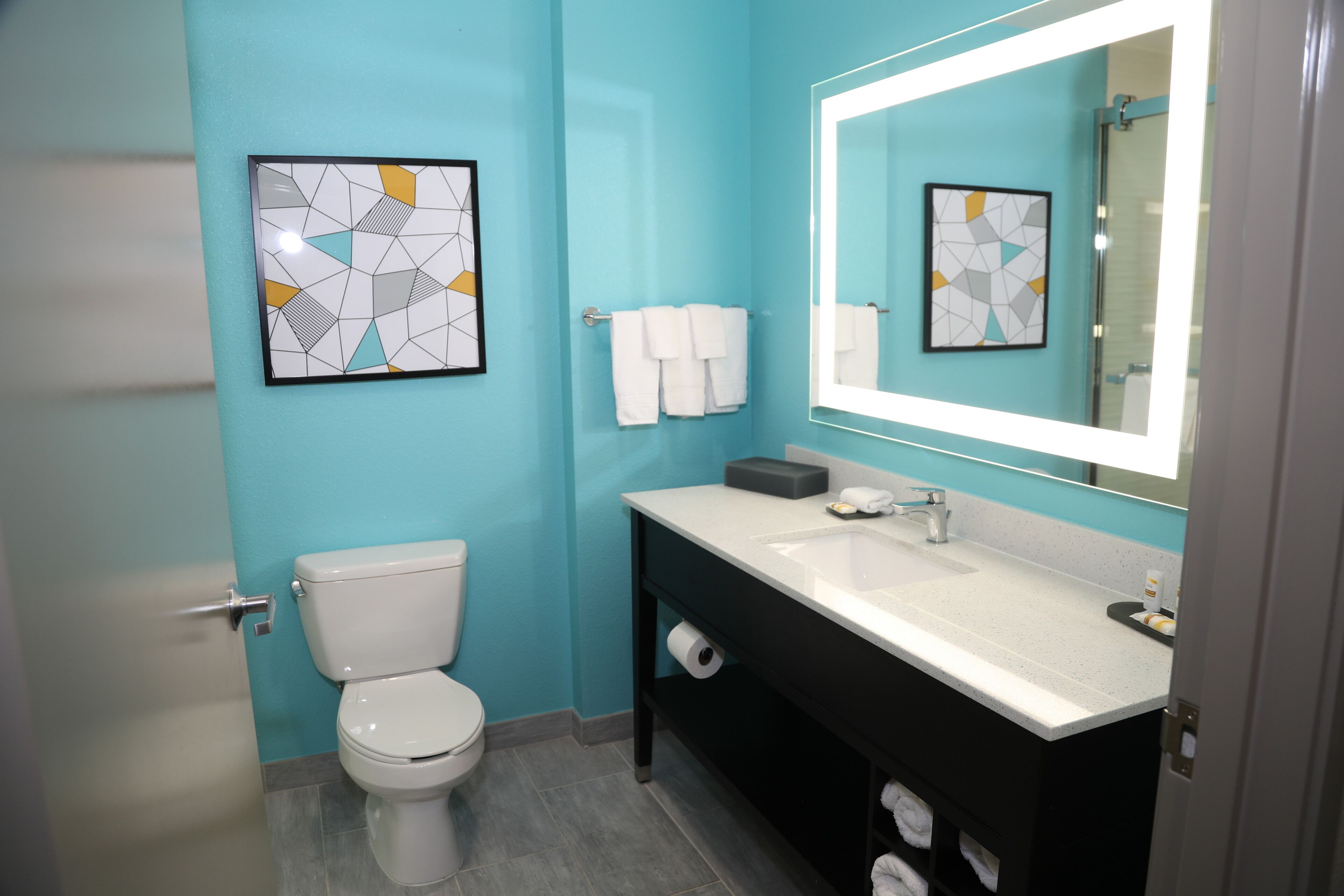 A modern bathroom with a toilet, sink, and mirror. There are towels hanging on the wall and a framed picture above the sink.