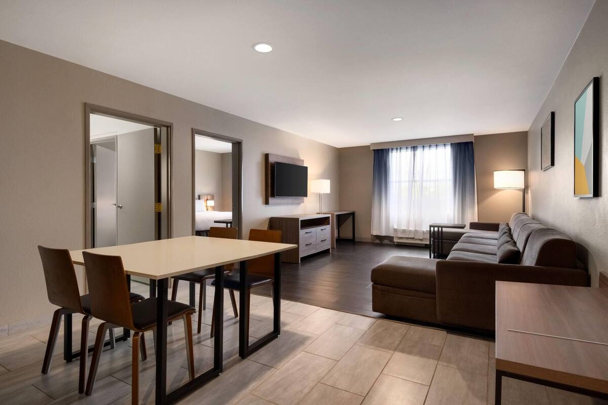 A modern hotel room with a dining area, a sofa, a TV, and a lamp.