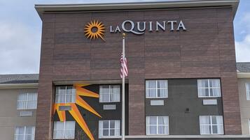 Um hotel La Quinta com design moderno, exibindo um sol proeminente no logotipo e um mastro com a bandeira americana.