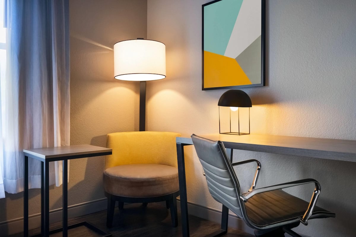 A hotel room with a modern chair, a small table, a lamp, and a wall-mounted abstract art piece.