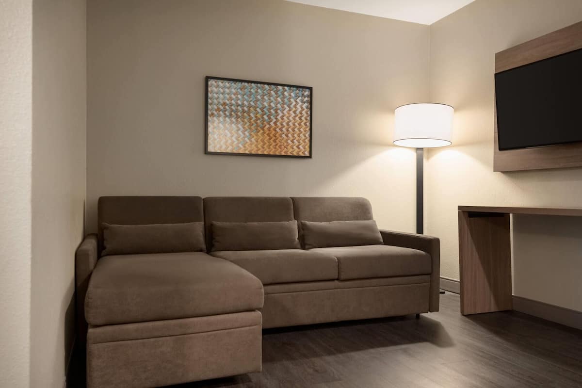 A modern hotel room with a brown sofa, a flat-screen TV mounted on the wall, and a floor lamp.