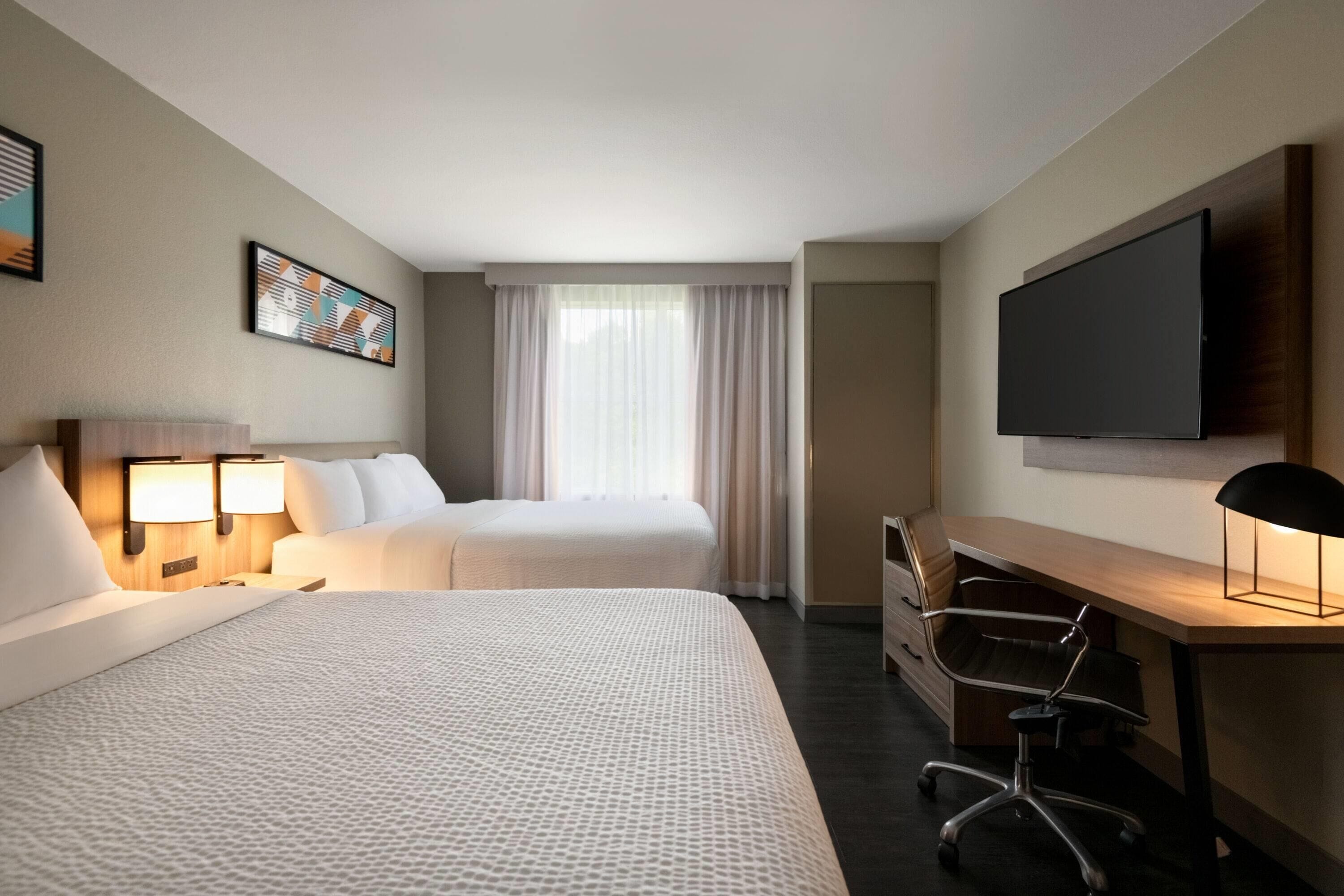 A hotel room with two beds, a desk, a chair, a TV, and a window with curtains.