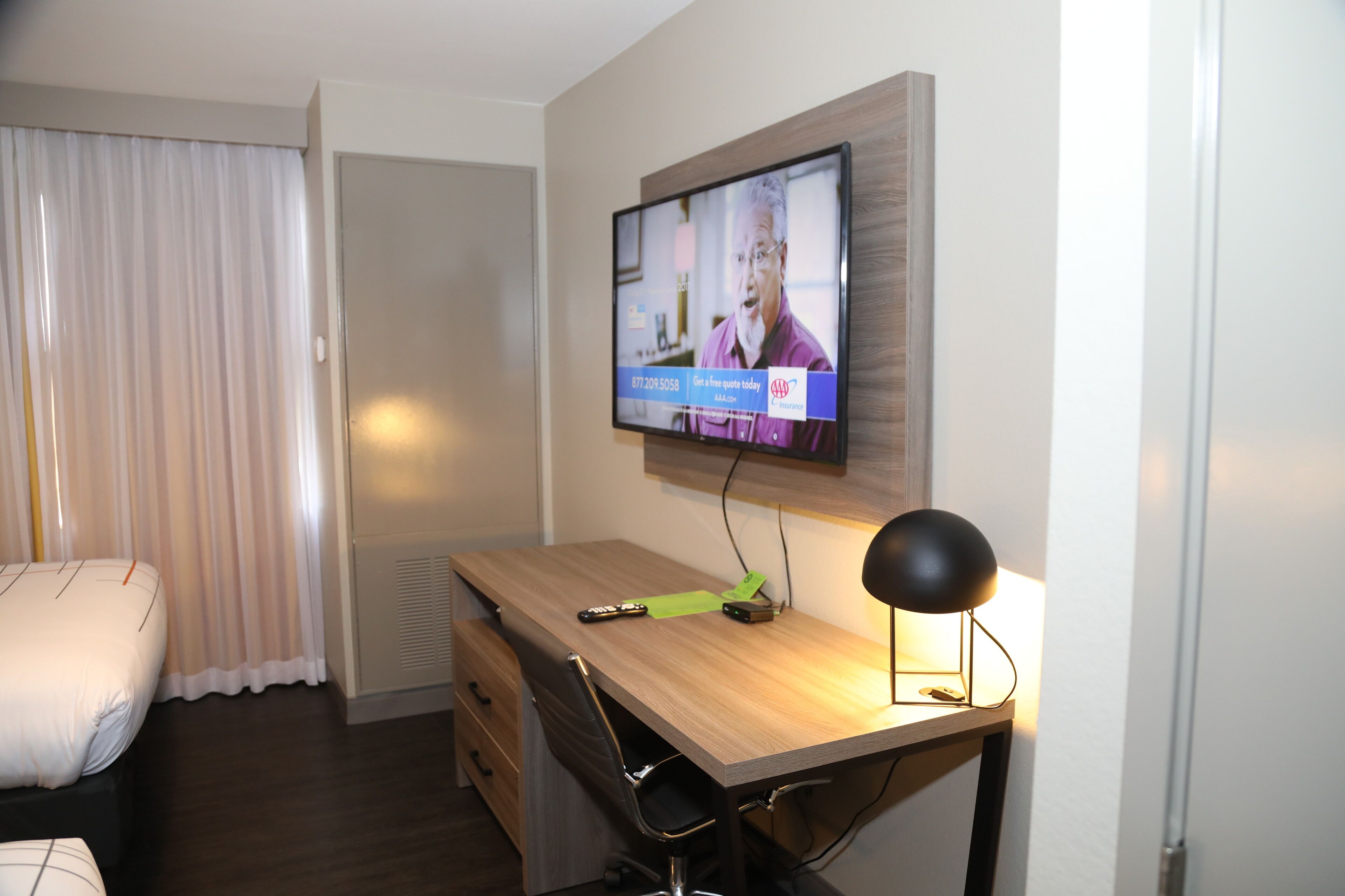 A hotel room with a wooden desk, a television mounted on the wall, a chair, and a bed with white bedding.