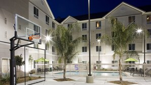 A pool area with a basketball hoop, palm trees, and a building with multiple windows and balconies. - Homewood Suites by Hilton  Fresno Airport/Clovis, CA (Clovis)