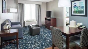 A hotel room with a sofa, a small table, a TV, a desk with chairs, a patterned rug, and a painting on the wall.