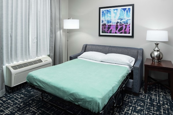 Una habitación de hotel con cama, mesita de noche, lámpara, aire acondicionado de pared y ventana con cortinas.