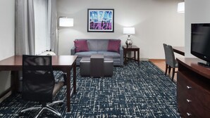 A hotel room with a desk, chair, sofa, ottoman, TV, lamp, and artwork on the wall. - Homewood Suites by Hilton  Fresno Airport/Clovis, CA (Clovis)