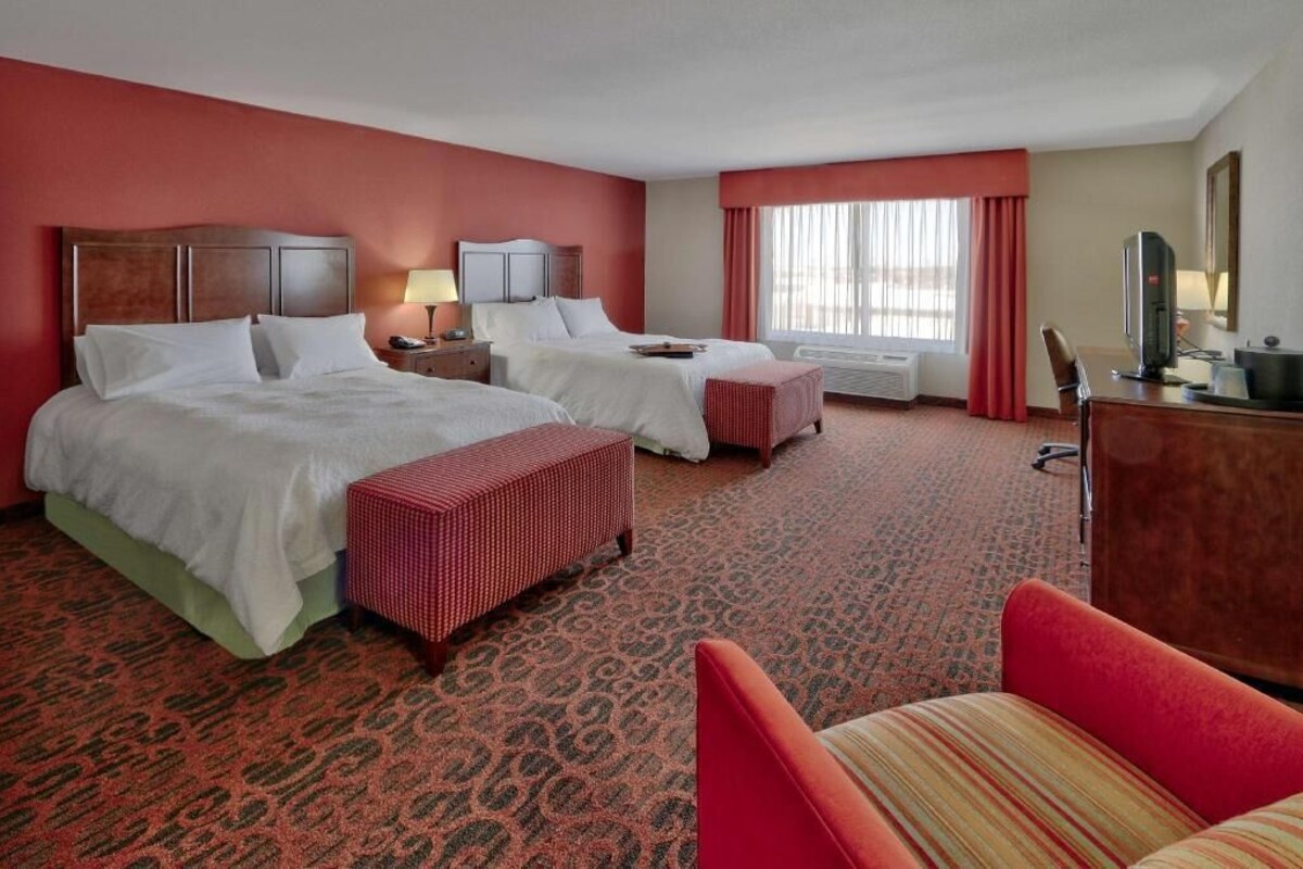 Quarto de hotel com duas camas, uma escrivaninha, uma cadeira, televisão e janela com cortinas.