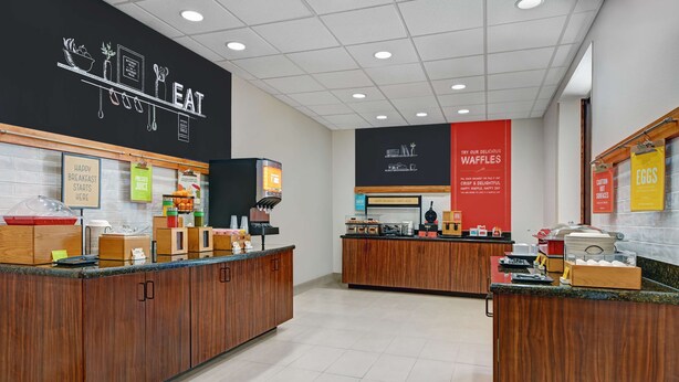 A modern cafe with wooden counters, a black wall with 'EAT' and food illustrations, and signs for waffles and eggs. - Hampton Inn Lordsburg (Lordsburg)