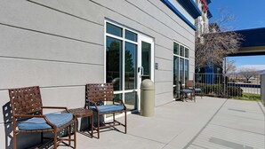 An outdoor seating area with two wooden chairs and a small table, a trash can, and a building entrance with a clear glass door. - Hampton Inn Lordsburg (Lordsburg)