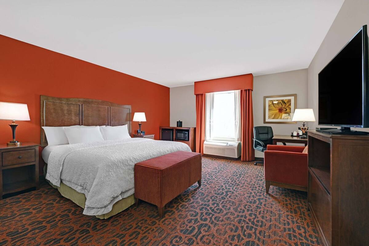 A hotel room with a bed, bedside tables, a desk, a chair, a TV, and a window with curtains.