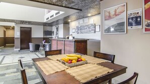Un comedor con una mesa de madera dispuesta con frutas y un mantel individual, una cocina al fondo y un techo con un diseño único.