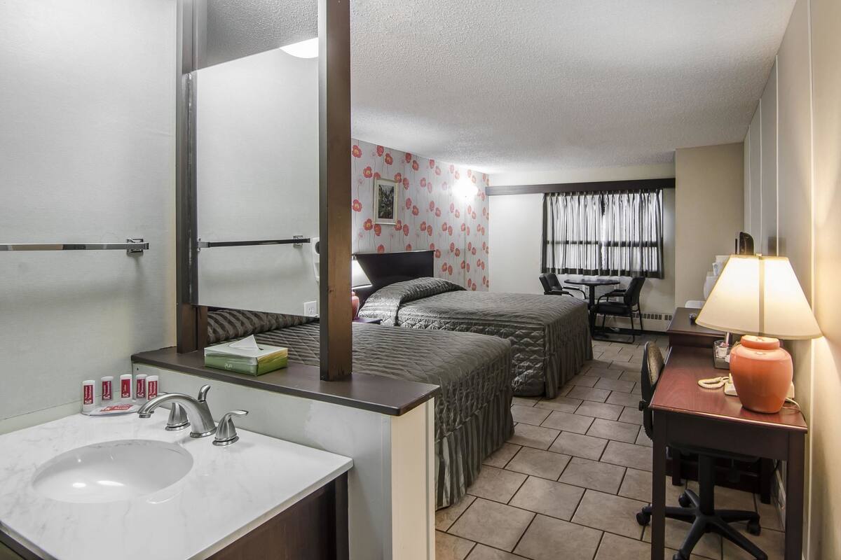 Une chambre d’hôtel avec un grand miroir, un lavabo, un lit avec deux oreillers, un bureau avec une lampe et une fenêtre avec des rideaux.