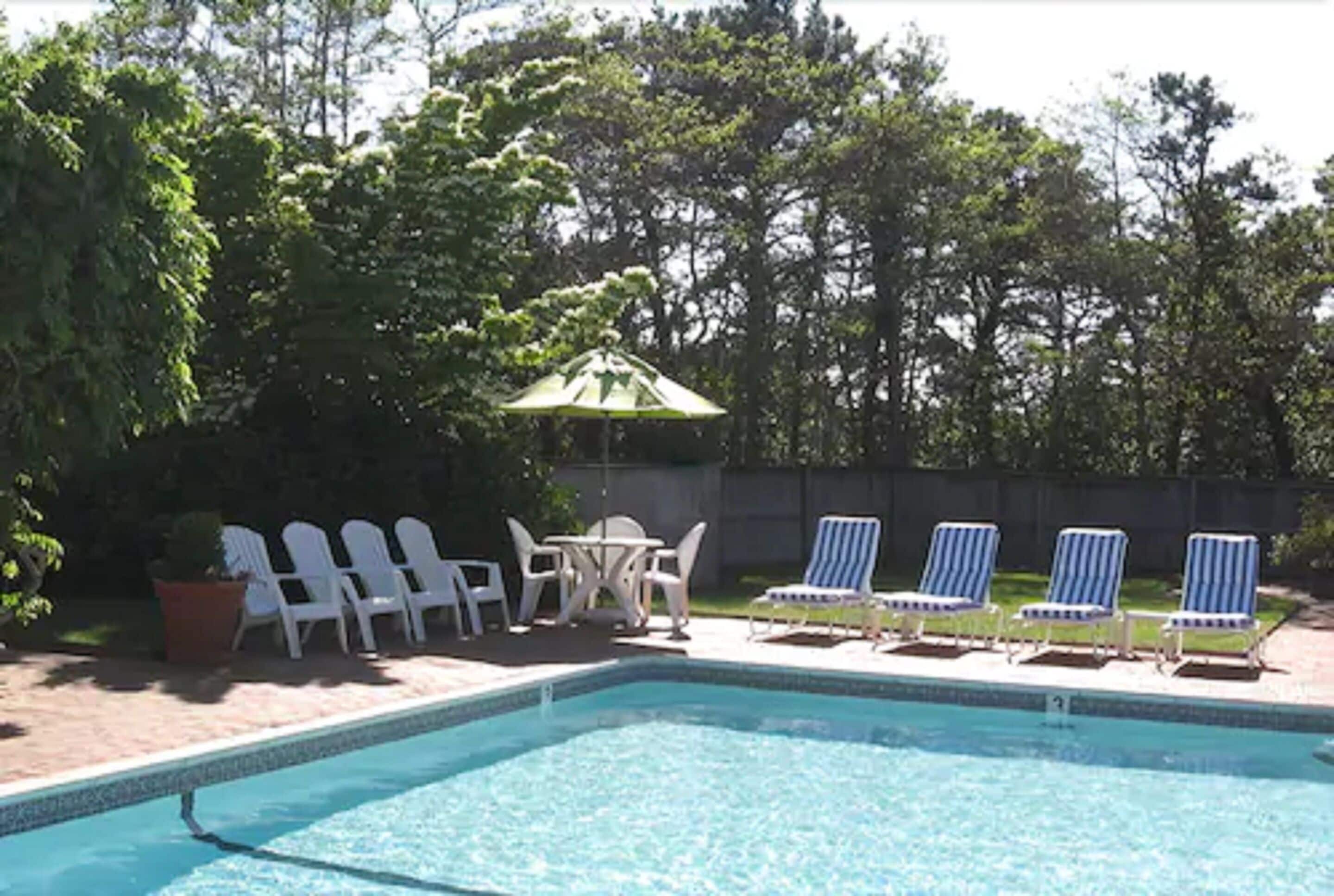 Una piscina rectangular con agua azul cristalina, rodeada de sillas reclinables blancas y una pequeña mesa con sillas.
