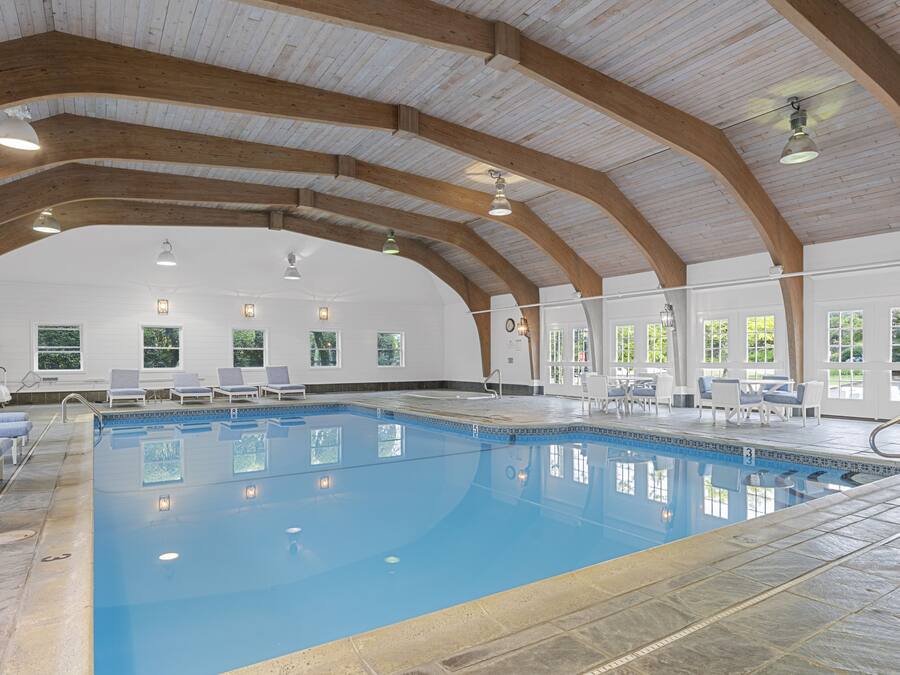 Une piscine intérieure dotée d’un haut plafond et de poutres en bois, entourée de chaises longues et de tables.