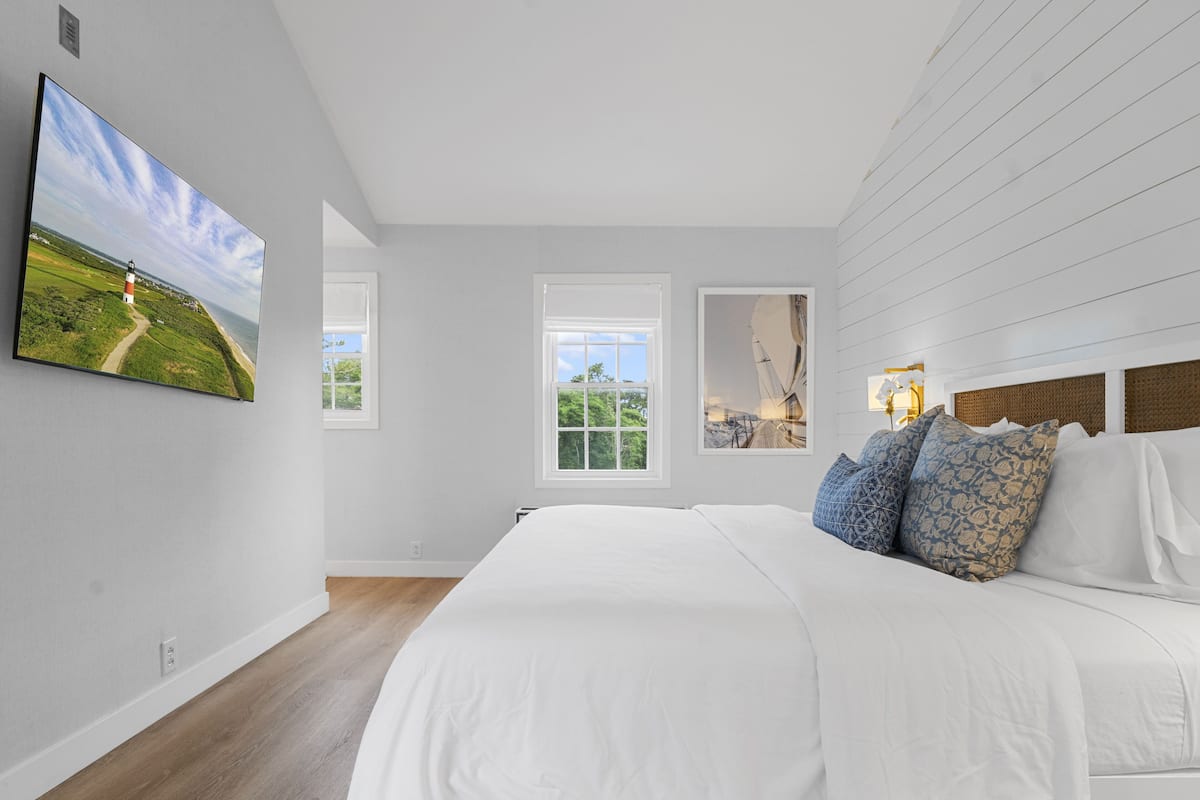 A bedroom with a large bed, a TV mounted on the wall, and two windows.