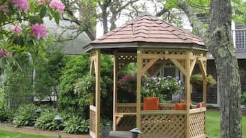 Un belvédère en bois, surmonté d’un toit en tuiles, entouré d’une végétation luxuriante et de plantes en fleurs.