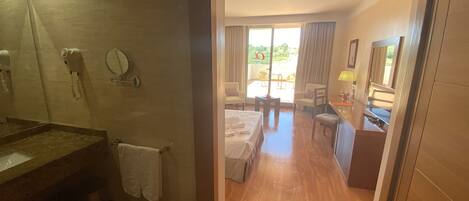 A hotel room with a wooden floor, a bed, a desk, a chair, a bathroom with a mirror, a hairdryer, and a towel rack.