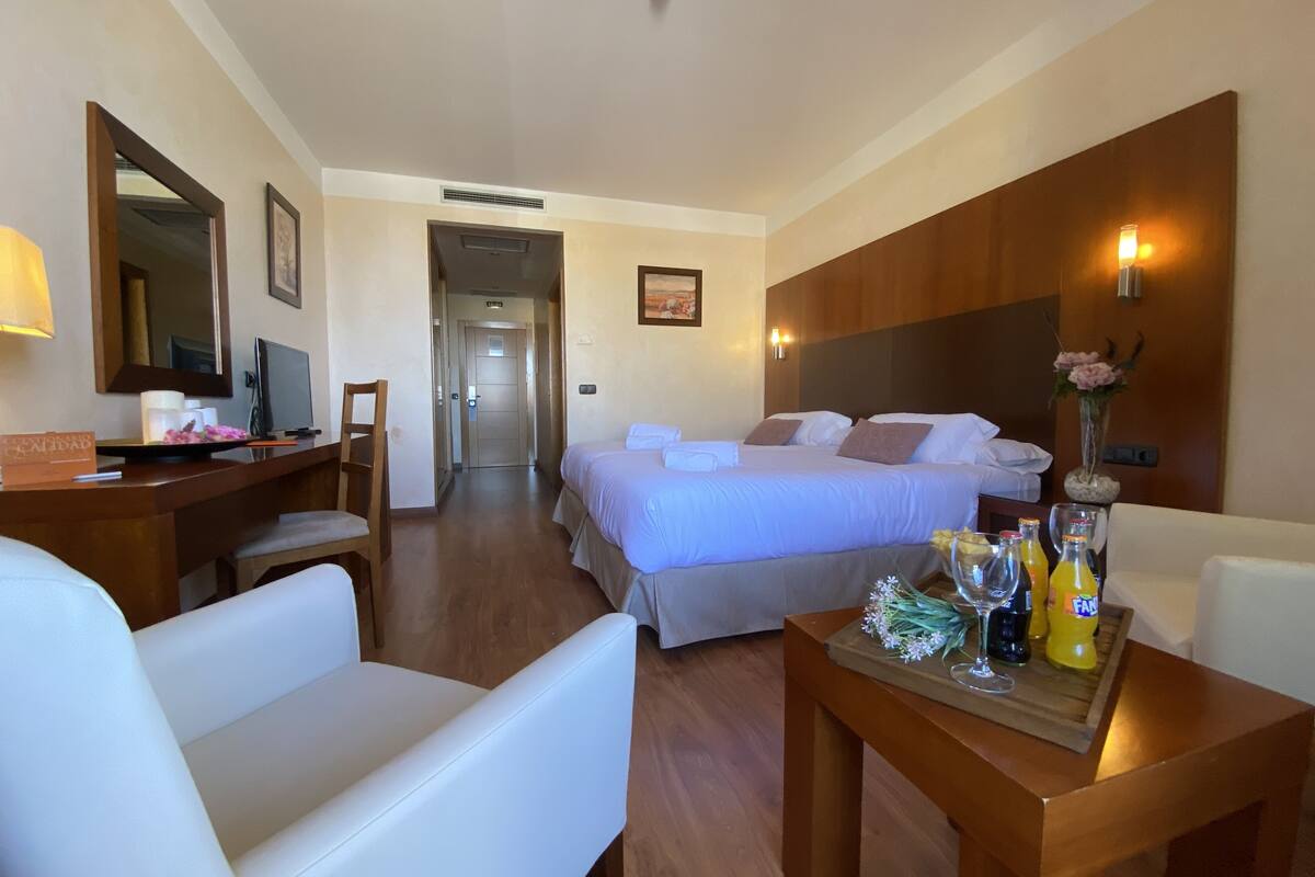 A hotel room with a bed, a desk with a computer, a chair, a table with drinks and flowers, and a mirror.