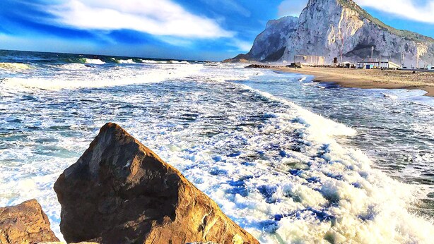 A coastal scene with a rocky foreground, waves crashing onto the shore, and a large cliff in the background. - Montera Plaza (Los Barrios)