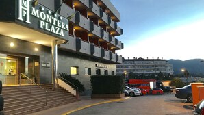 A multi-story hotel with a sign reading "Monteria Plaza" and "Restaurante" indicating a restaurant on the ground floor. There are steps leading to the entrance, a parking area with cars, and a view of the surrounding buildings and mountains in the background. - Montera Plaza (Los Barrios)