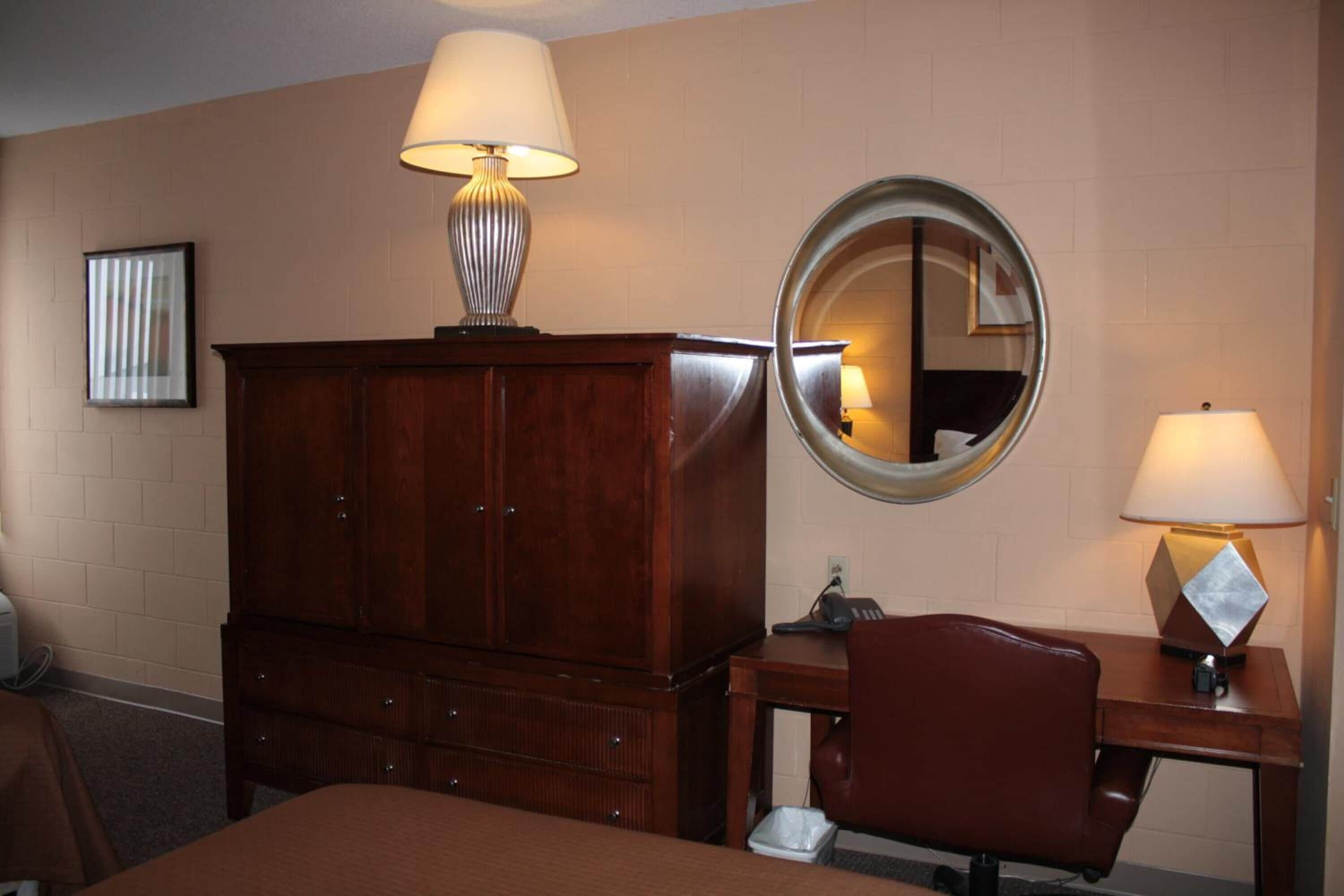 Una habitación de hotel con un aparador de madera, un espejo redondo, un escritorio con lámpara y una cama.