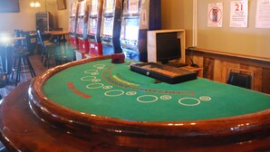 A casino table with a green felt surface, surrounded by chairs and a bar area with slot machines. - Sunlac Inn (Lakota)