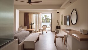 Een moderne hotelkamer met een bed, een bureau, een stoel, een bankje, een plafondventilator en een balkon met gordijnen.