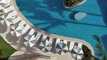 An aerial view of a pool area with sun loungers and umbrellas, surrounded by a stone deck and palm trees.