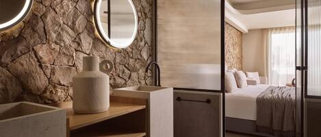 A modern bathroom with a stone wall, a wooden vanity, a large mirror, and a bed in the adjacent room.